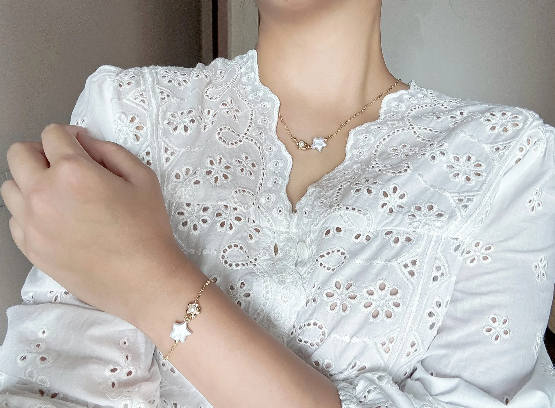 Model in a white eyelet-embroidered blouse wearing the star and flower necklace, with a matching fine-chain bracelet on the wrist.