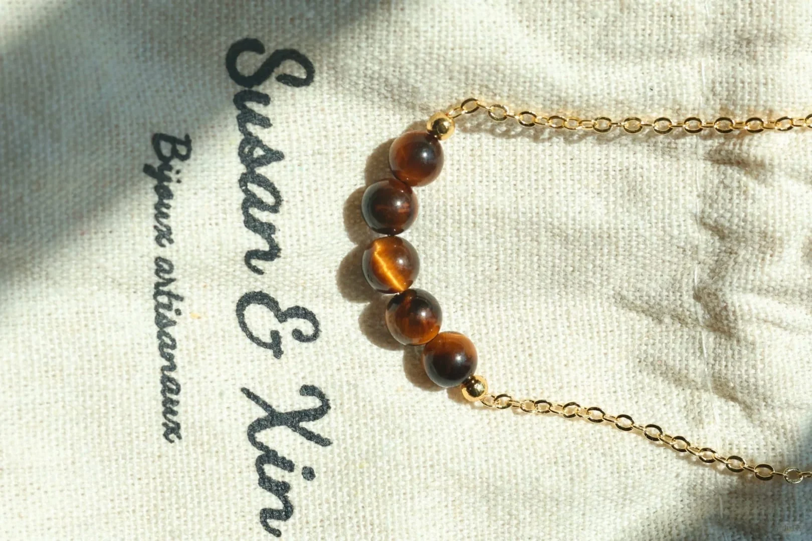 Close-up of a fine gold-toned chain necklace with five round tiger’s eye beads in a row at the centre, flanked by small gold-toned beads, on a Susan&Xin fabric pouch.