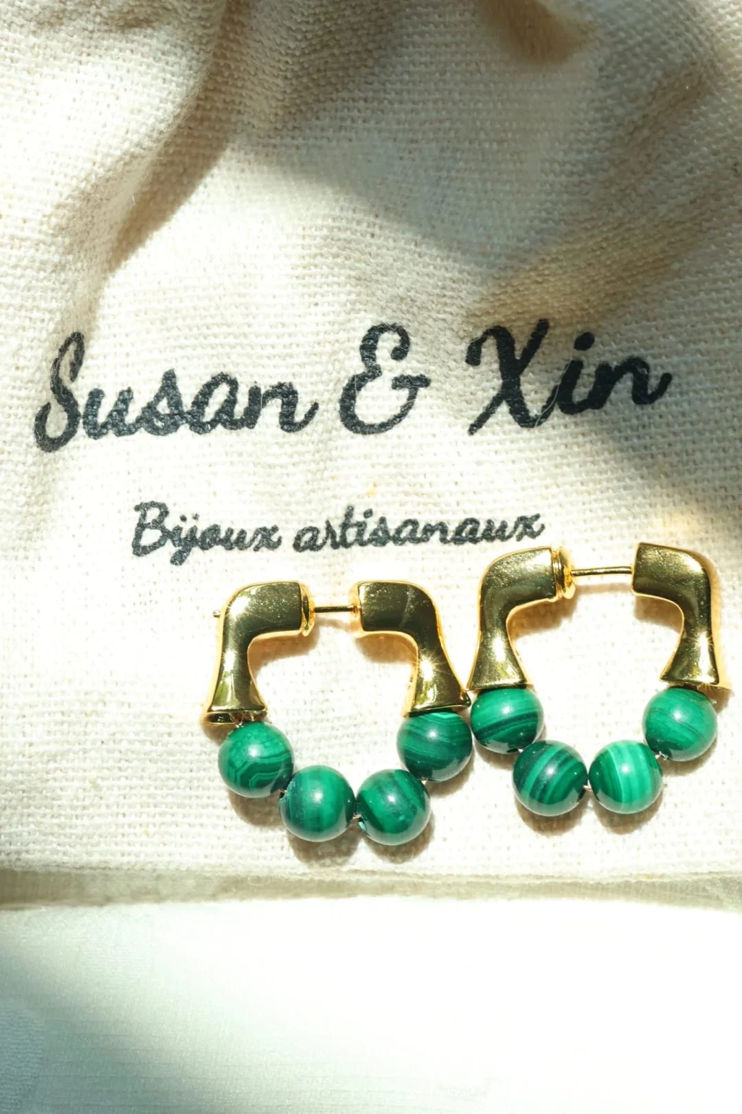 Pair of gold-toned hoop earrings with a squared upper section and four banded malachite beads along the bottom of each hoop, on a Susan&Xin fabric pouch.