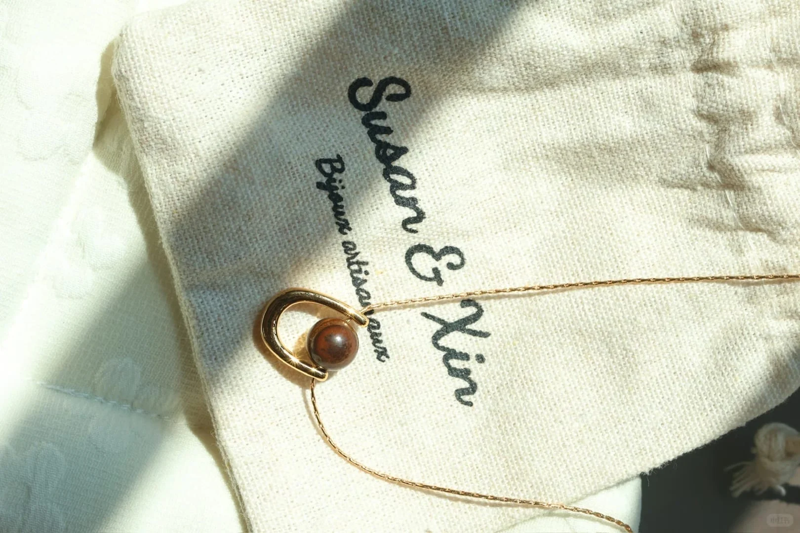 Very fine gold-toned chain necklace with an open arc-shaped pendant holding a single deep-brown tiger’s eye bead, on a Susan&Xin fabric pouch.