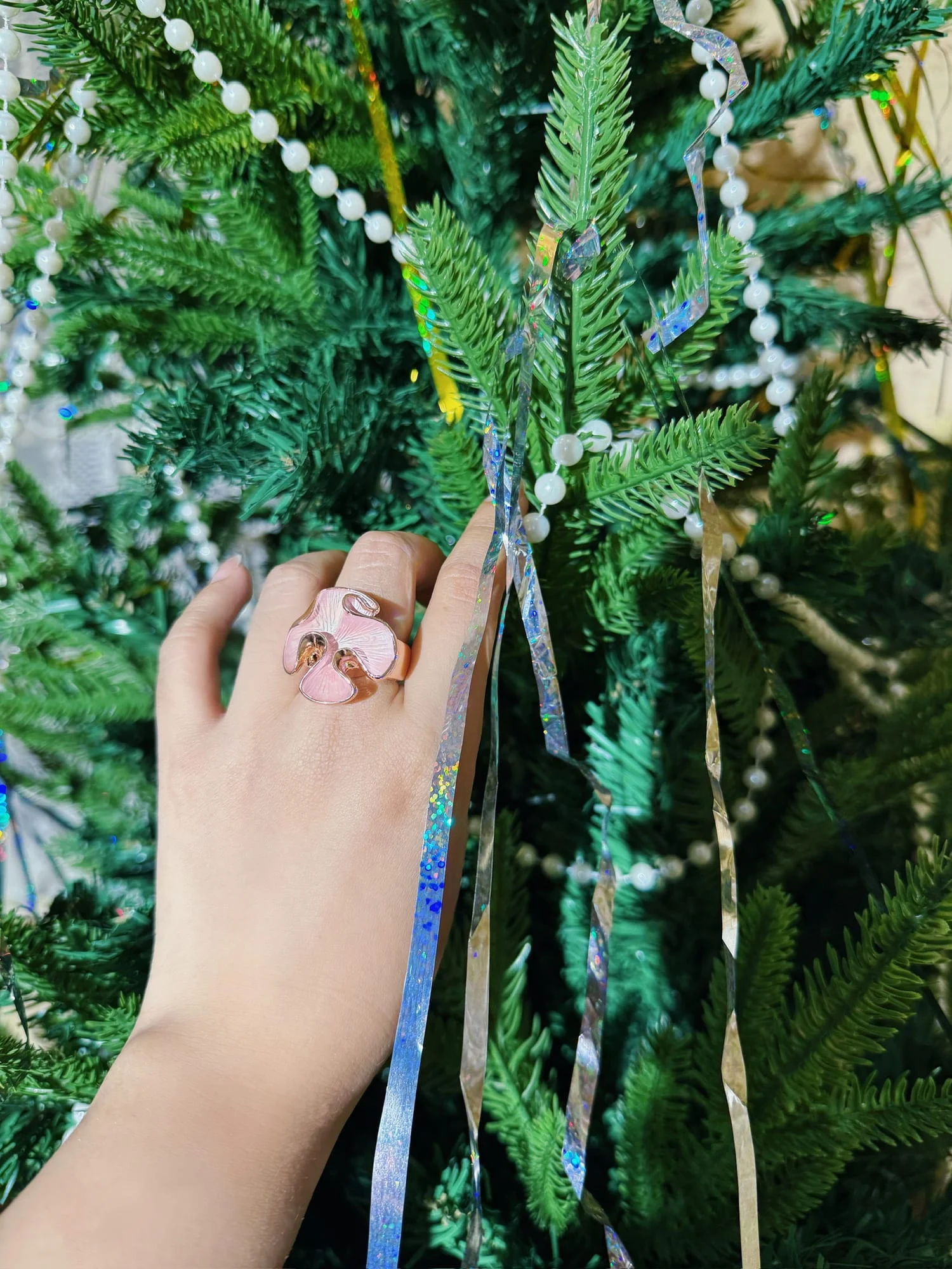 Hand wearing the pink enamel orchid ring, reaching into a Christmas tree hung with bead garlands and tinsel.